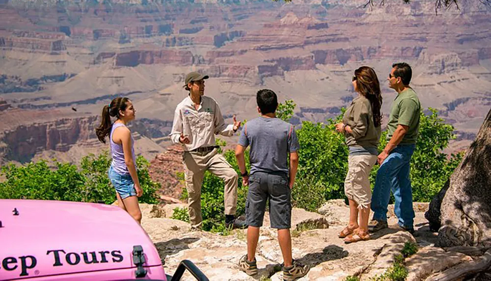 A group of tourists is engaging with a guide near the edge of a canyon with a pink Jeep Tours vehicle visible in the foreground