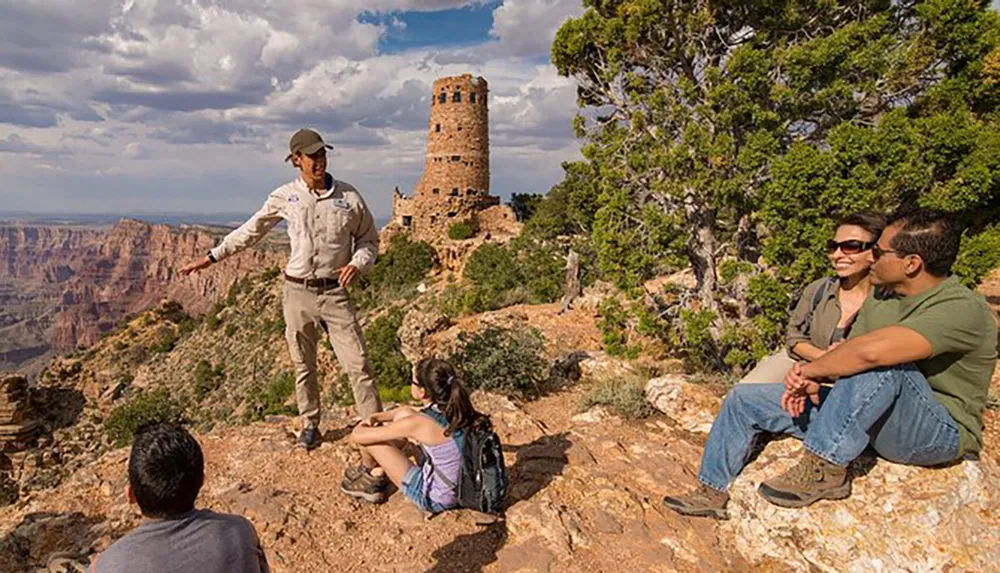A park ranger is giving a tour to visitors near the edge of the Grand Canyon with a stone watchtower in the background