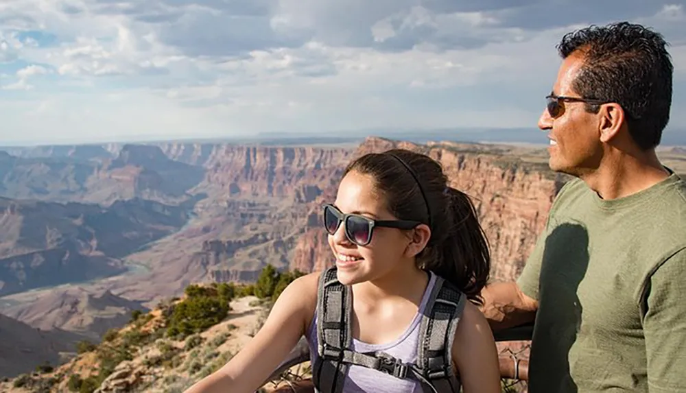 A man and a young girl with sunglasses are enjoying a scenic view of the Grand Canyon under a partly cloudy sky