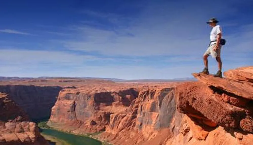 A person stands on the edge of a steep cliff above a canyon with a river meandering below under a clear blue sky
