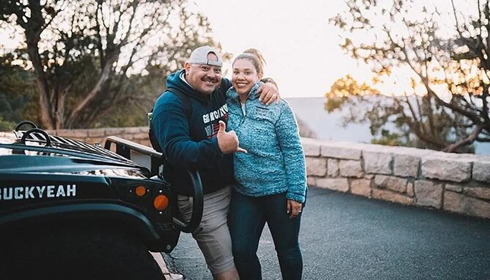 A happy couple is posing for a photo next to an off-road vehicle with a license plate reading FUCKYEAH all against a backdrop of trees and a sunset