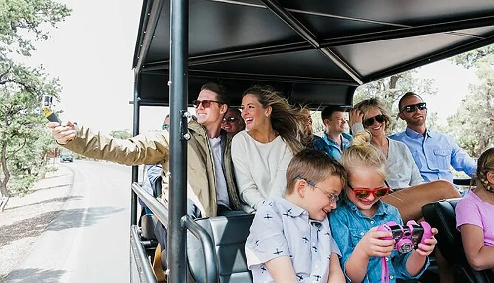 A group of smiling people including children is enjoying a ride on an open-air tour vehicle with a man taking a selfie at the front