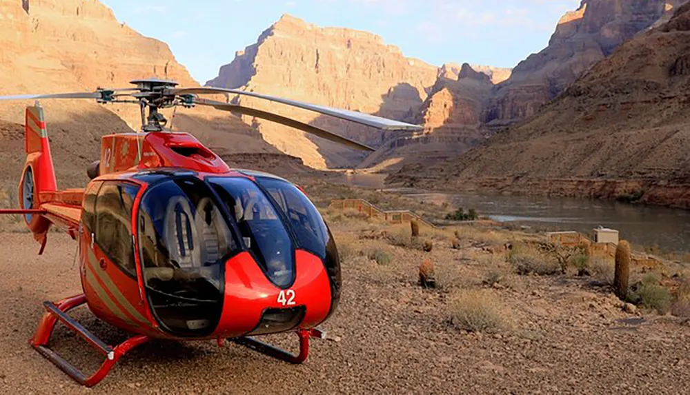 A red helicopter is parked on a dusty terrain with a river and towering canyon walls in the background