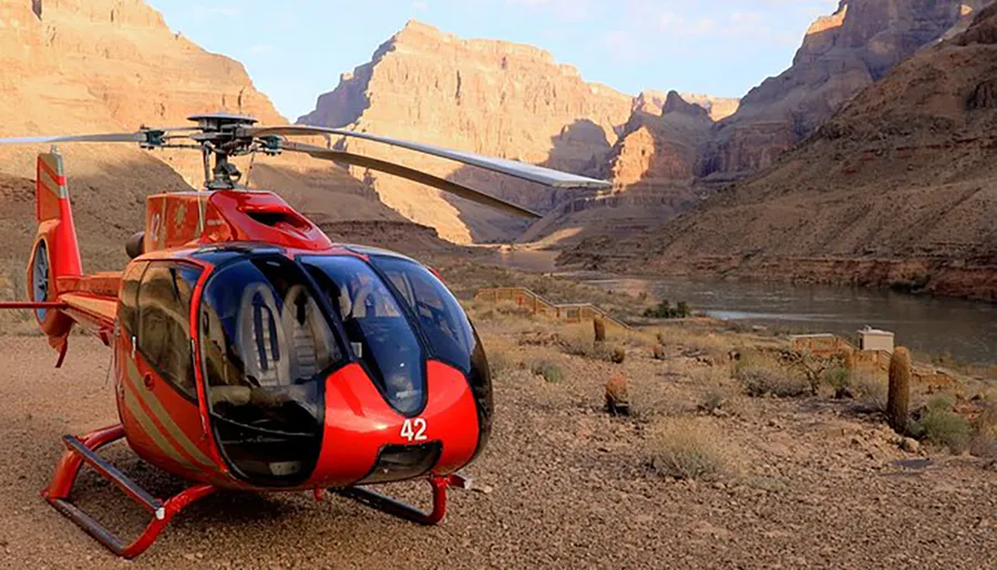 A red helicopter is parked on a dusty terrain with a river and towering canyon walls in the background.