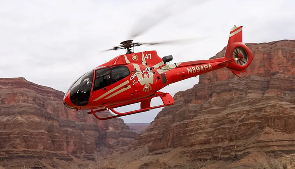 A red helicopter with the words Grand Canyon is flying in front of a rugged cliff indicating it may be on a sightseeing tour or conducting operations within the Grand Canyon area
