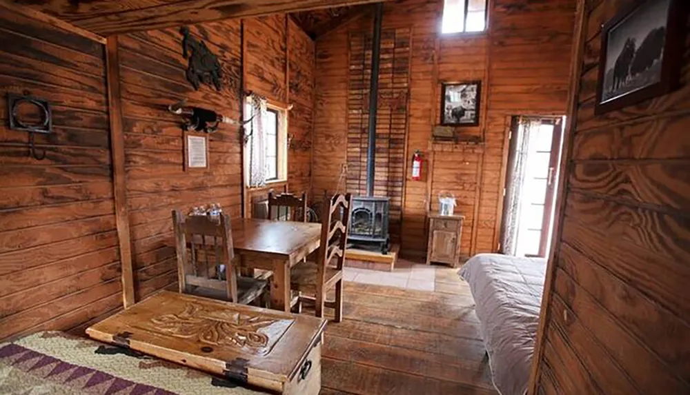 The image shows the rustic interior of a cabin featuring wood-paneled walls and ceilings a dining area with simple wooden furniture a cozy stove and a bed in the corner near a door allowing natural light in