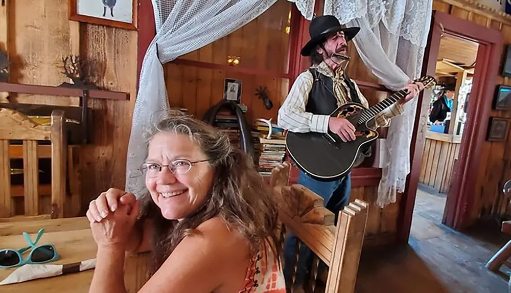 A woman smiles at the camera while a man in a cowboy hat plays the guitar in the background of what appears to be a rustic-themed room