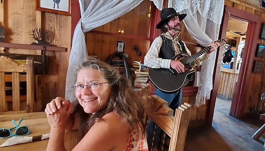 A woman smiles at the camera while a man in a cowboy hat plays the guitar in the background of what appears to be a rustic-themed room.