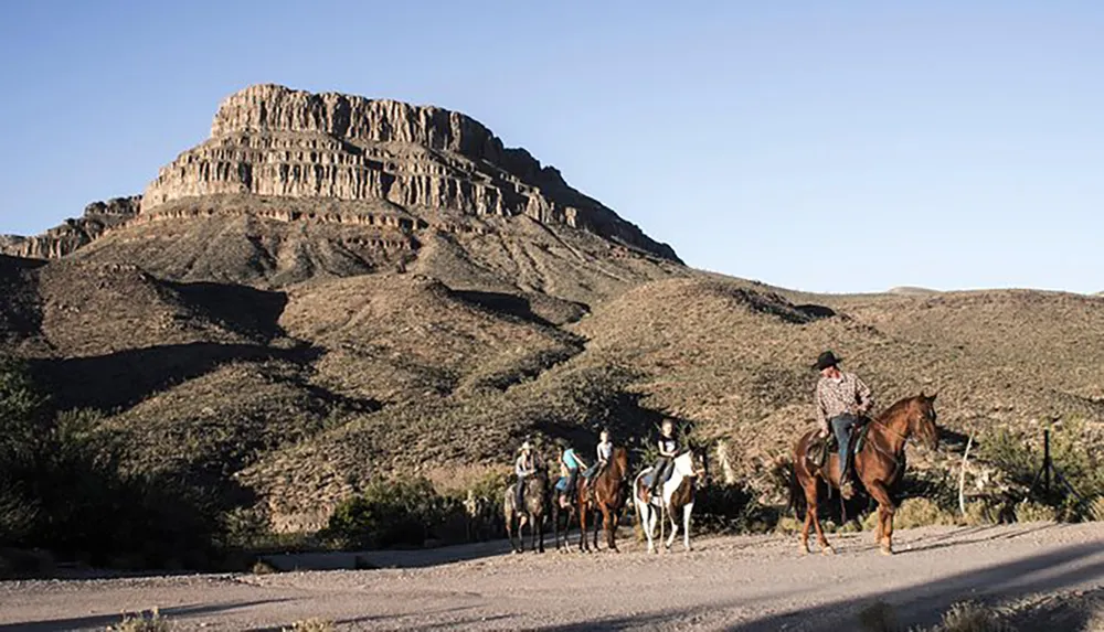A group of riders on horseback are traveling on a dirt road with a large layered rocky hill in the background under a clear sky