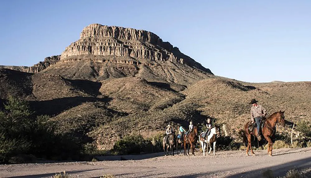 A group of horseback riders travels along a dirt road with a large stratified mesa rising in the background under a clear sky