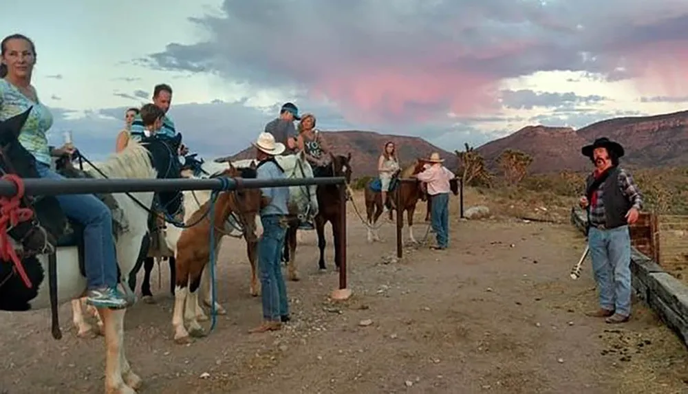 A group of people wearing cowboy hats are with horses at a fence line against a backdrop of a mountainous landscape under a pink-hued sky