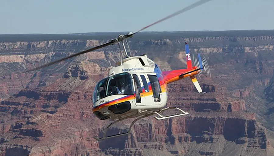 A helicopter is flying in front of the expansive and layered rock formations of the Grand Canyon under a clear blue sky.