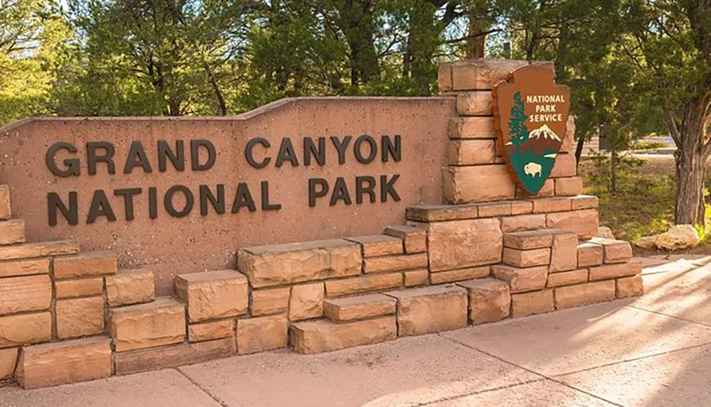 The image shows the entrance sign to Grand Canyon National Park featuring large metal letters on a stone backdrop with a National Park Service emblem to the side