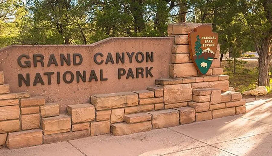 The image shows the entrance sign to Grand Canyon National Park, featuring large metal letters on a stone backdrop with a National Park Service emblem to the side.