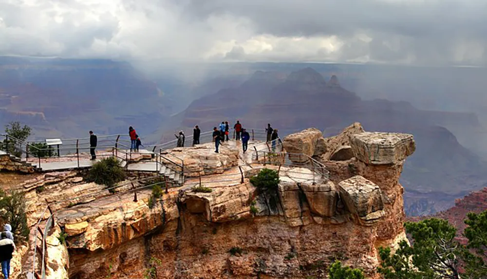 Tourists are exploring a viewpoint at the Grand Canyon with safety railings against a backdrop of vast cliffs and canyons under a cloudy sky