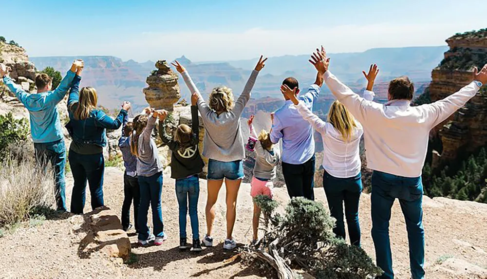 A group of people is standing with their backs to the camera raising their arms in joy in front of a vast canyon landscape