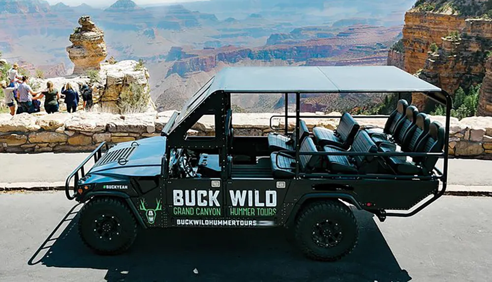 A tour vehicle labeled BUCK WILD Grand Canyon Hummer Tours is parked at a viewing area where people are enjoying the scenic backdrop of the Grand Canyon