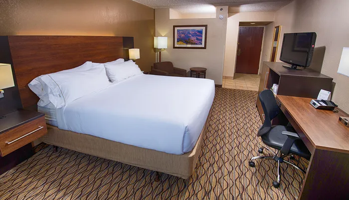 The image shows a neatly arranged hotel room with a king-sized bed a work desk with a chair and a flat-screen TV