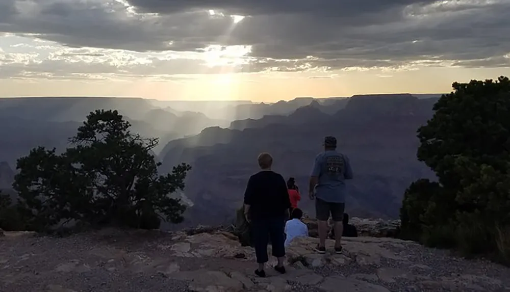 A group of people enjoys a sunset at the edge of a vast canyon likely the Grand Canyon with layered rock formations fading into the distance under a partly cloudy sky