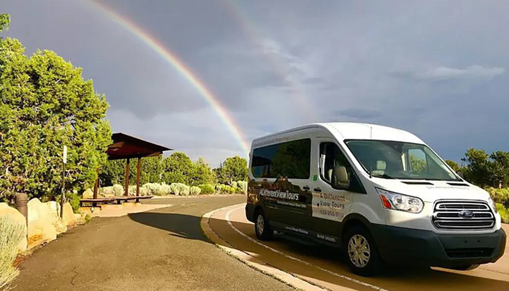 A tour van is parked along a road with a vibrant rainbow in the background under a stormy sky