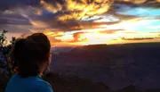 A person gazes out at a breathtaking sunset over the vast canyons painted with shades of orange and purple in the sky.