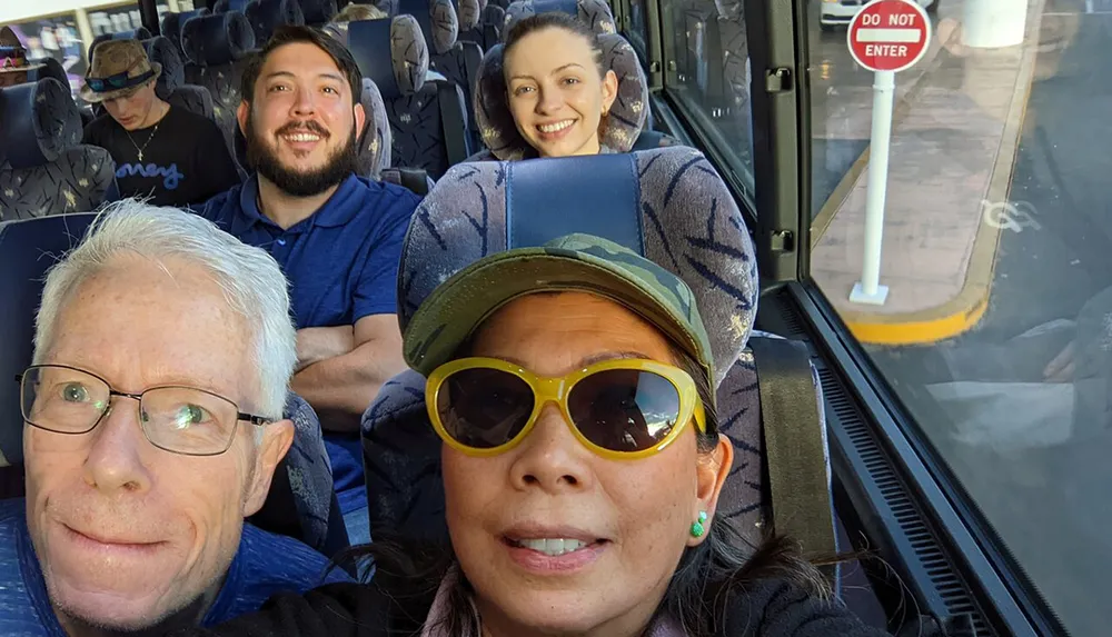 Four individuals are posing for a group selfie on a bus with one capturing the front-facing shot and the others smiling behind them