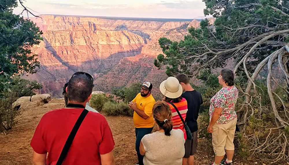 A group of tourists are attentively listening to a guide while standing at an overlook with a view of the Grand Canyon during sunset