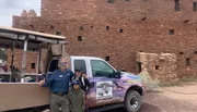 Two adults and a child pose for a photo in front of a Grand Canyon tour vehicle, with an ancient-looking stone building in the background.