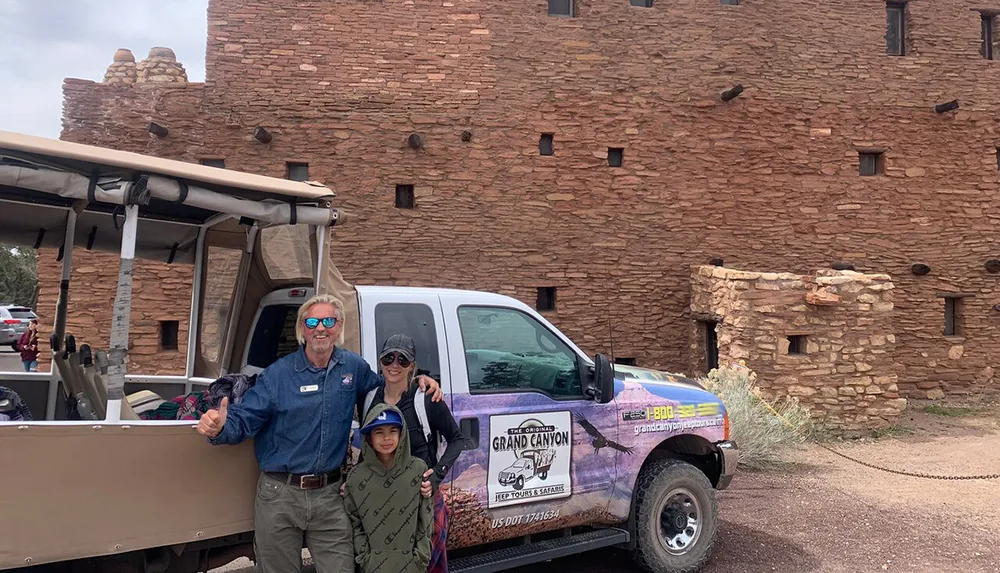 Two adults and a child pose for a photo in front of a Grand Canyon tour vehicle with an ancient-looking stone building in the background