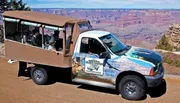 A group of tourists is riding in a vehicle with a Grand Canyon-themed wrap design, touring near the edge of the canyon.