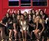 A group of people in swimwear is posing cheerfully beside a red bus with the slogan Life is a Party and the iconic Route 66 sign indicating a fun social outing probably linked to a bachelorette celebration as indicated by one person wearing a Bride swimsuit