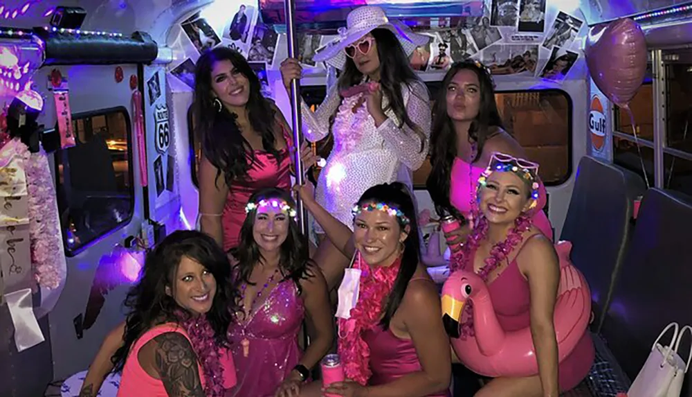 A group of women dressed in pink-themed party attire are posing joyfully inside a festively decorated bus with a flamingo inflatable and party lights