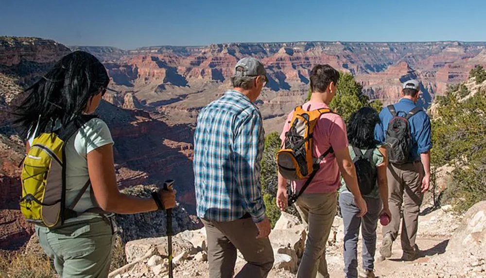 A group of hikers with backpacks is walking towards the edge of the Grand Canyon under a clear blue sky