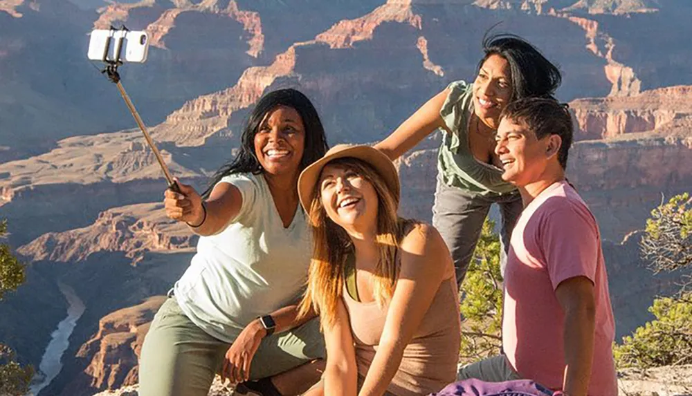 Four friends are taking a group selfie with a smartphone on a selfie stick with a scenic canyon backdrop illuminated by sunlight