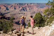 Two hikers are taking a selfie with the expansive view of the Grand Canyon in the background.