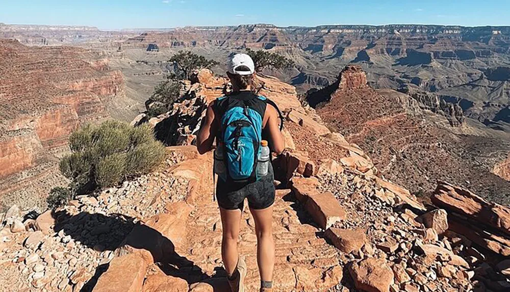 A hiker with a backpack is standing on a rocky trail looking out over the vast expanse of the Grand Canyon