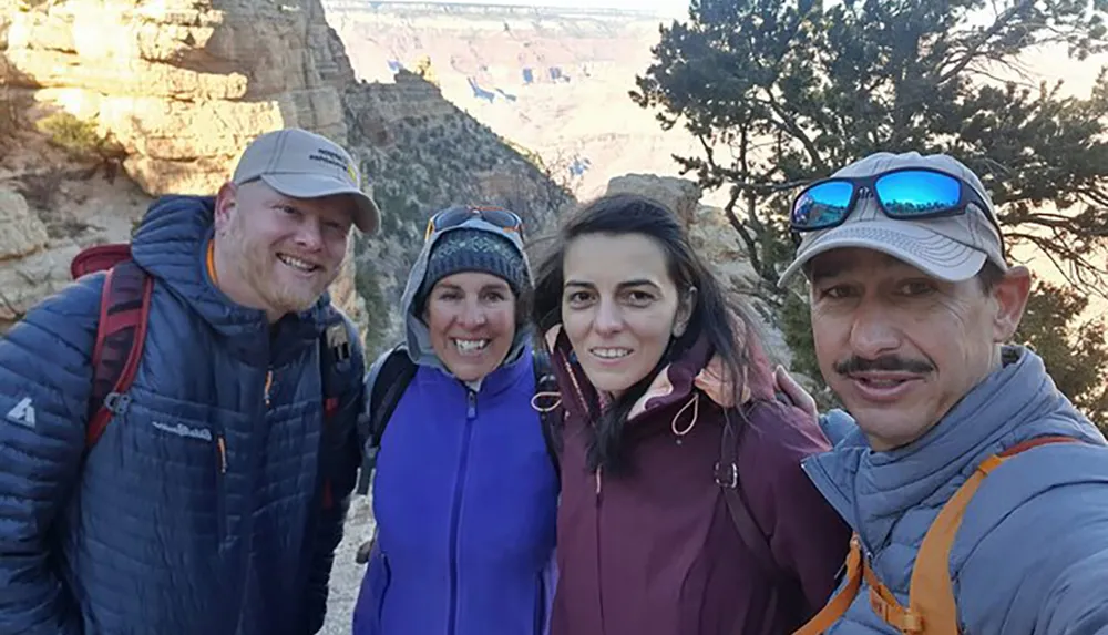 Four smiling hikers are taking a group selfie with a scenic canyon backdrop