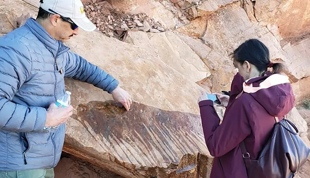 Two people are examining a large rock with petroglyphs on it with one person taking a photo of the engravings