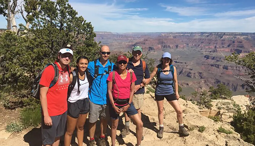 A group of six hikers is posing for a photo in front of the Grand Canyon on a sunny day