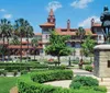 The image shows a formal garden with manicured hedges and a statue in the foreground set against the backdrop of a historic building with red-tiled roofs and towers