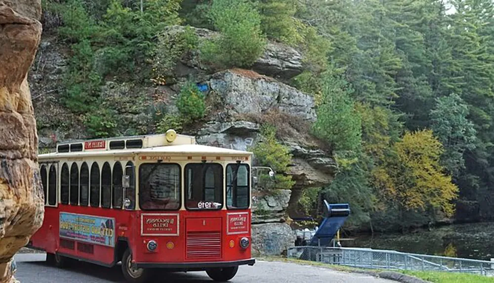 A red trolley bus with advertisements on its side is driving by a rocky outcrop surrounded by lush greenery and a few trees with yellow autumn leaves