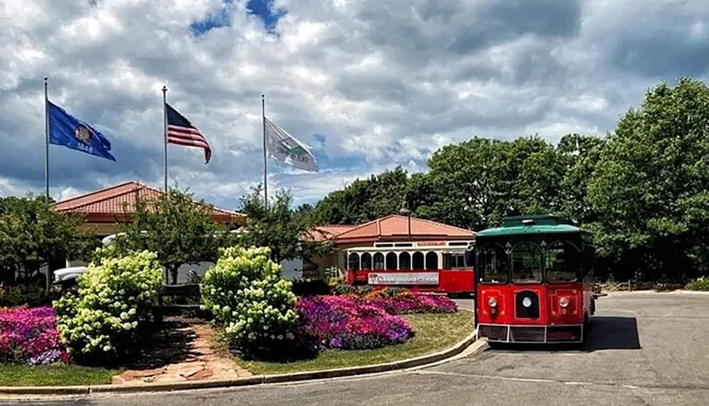 A red trolley car is parked in front of a building with a terracotta roof surrounded by lush landscaping and flying flags under a partly cloudy sky