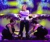 A performer is standing onstage with mechanical wings extended while two assistants appear to be adjusting the base of the wings amidst colorful stage lighting