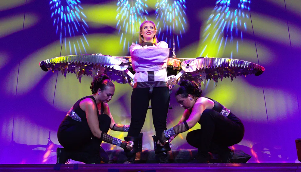 A performer is standing onstage with mechanical wings extended while two assistants appear to be adjusting the base of the wings amidst colorful stage lighting
