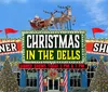 This image features a festive stage performance with a person dressed as Santa Claus dancing among performers in toy soldier costumes under a banner encouraging people to visit Santas Workshop for the Most Fun in the Dells at Christmas