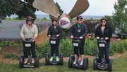 Four people wearing helmets are standing in a line on Segways in front of a large airplane propeller sculpture.