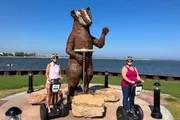 Two people on Segways are posing next to a large metal statue of a bear by a lakeside.