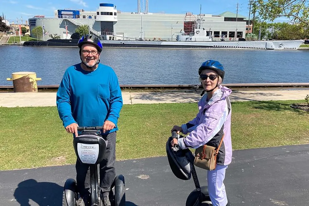 Two people wearing helmets are smiling for a photo while standing with Segways near a waterfront with a ship in the background