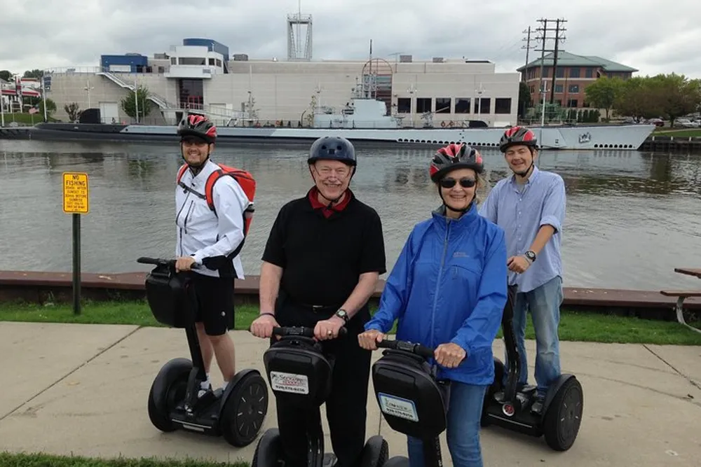 Four people wearing helmets are standing with Segways by a riverside with a boat and buildings in the background