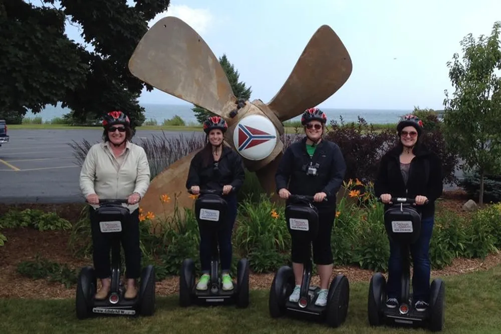 Four individuals wearing helmets are standing on Segways in front of a large propeller monument
